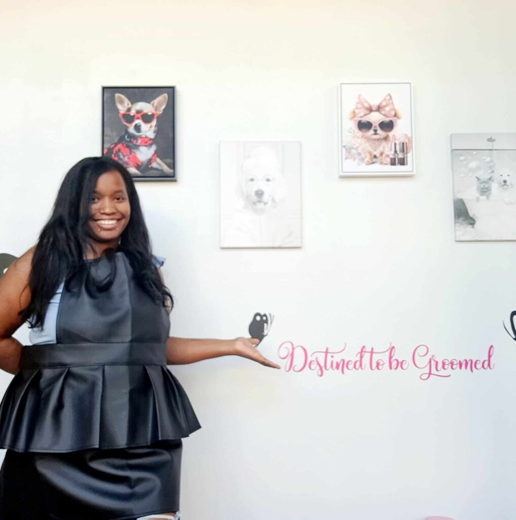 A woman in a black dress stands smiling and gesturing toward a wall with framed dog photos and the words Destined to be Groomed in pink script.